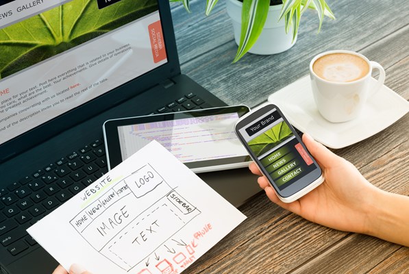 Hand holding a mobile on a work desk that has a laptop and tablet