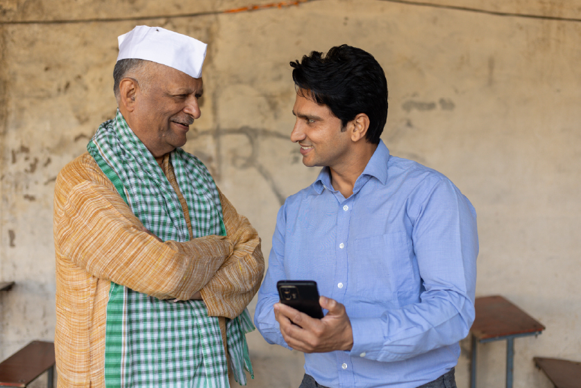 Young Indian bank person or finance adviser showing information to senior farmer on smartphone